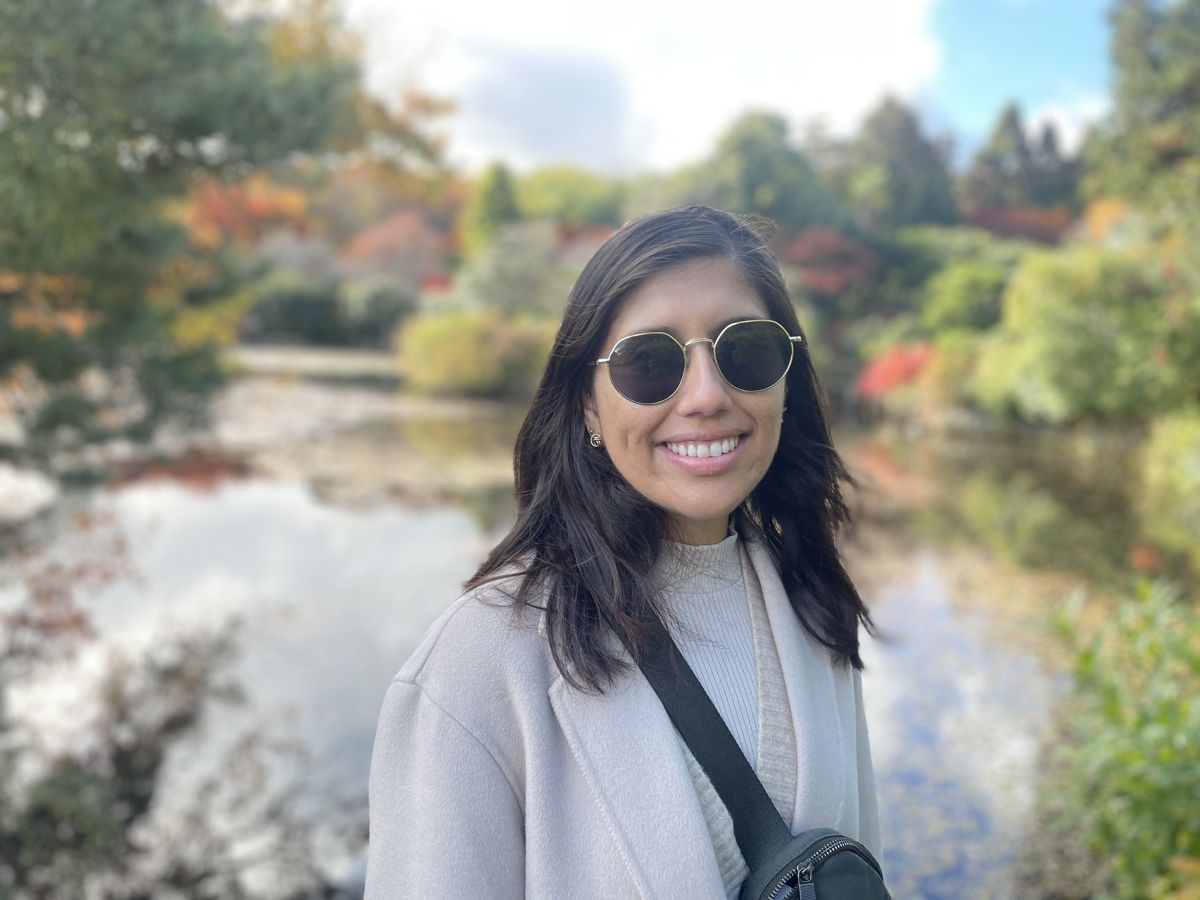 Priyanka smiling outdoors beside a pond with autumn trees